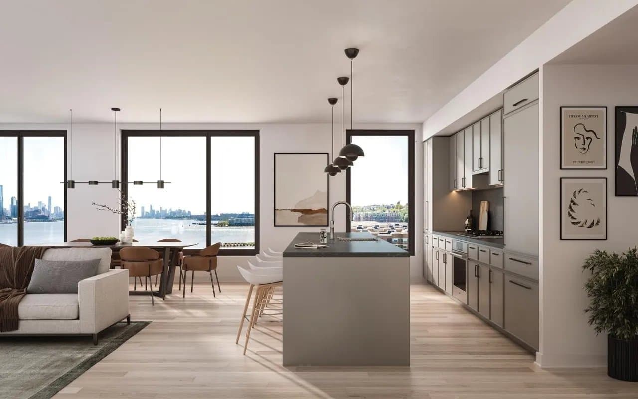 An open-plan condo kitchen and living room with tall windows looking across the Hudson River to the Manhattan skyline