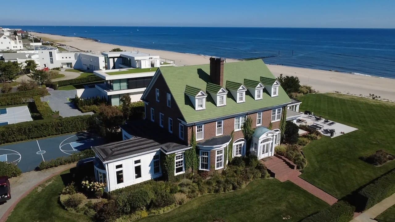 Drone view of a brick colonial with a green roof and dormers on a wide lawn beside a New Jersey beach
